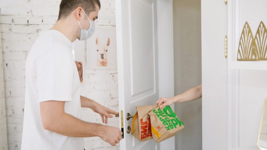 A man in a face mask receives a contactless food delivery at his door, emphasizing pandemic safety.